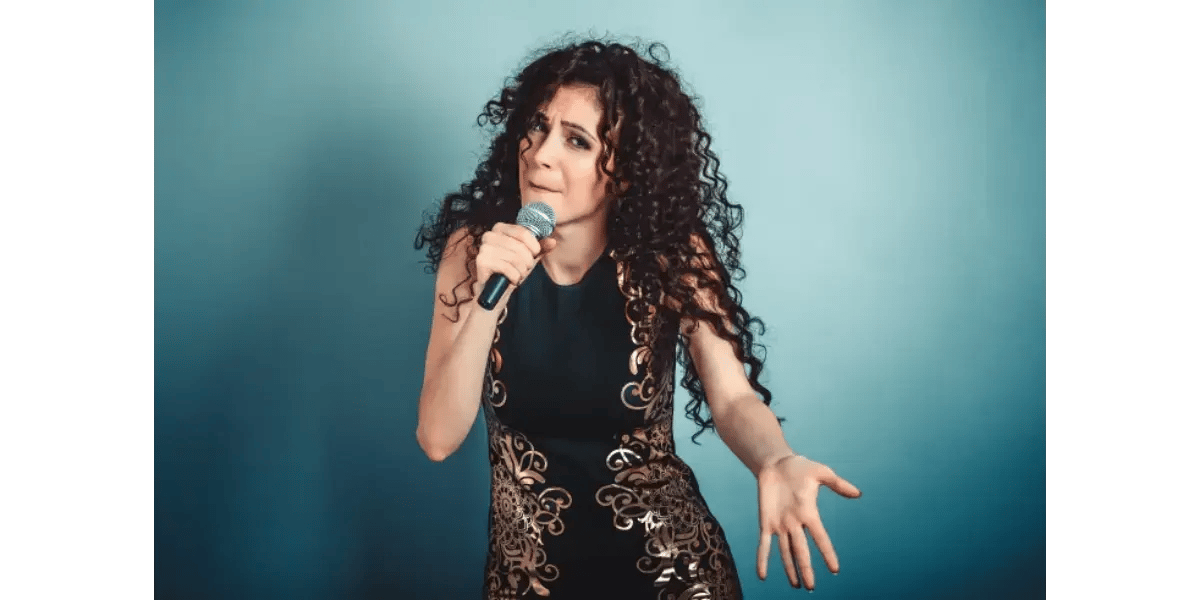 A woman with dark curly hair singing passionately into a microphone against a vibrant blue background.