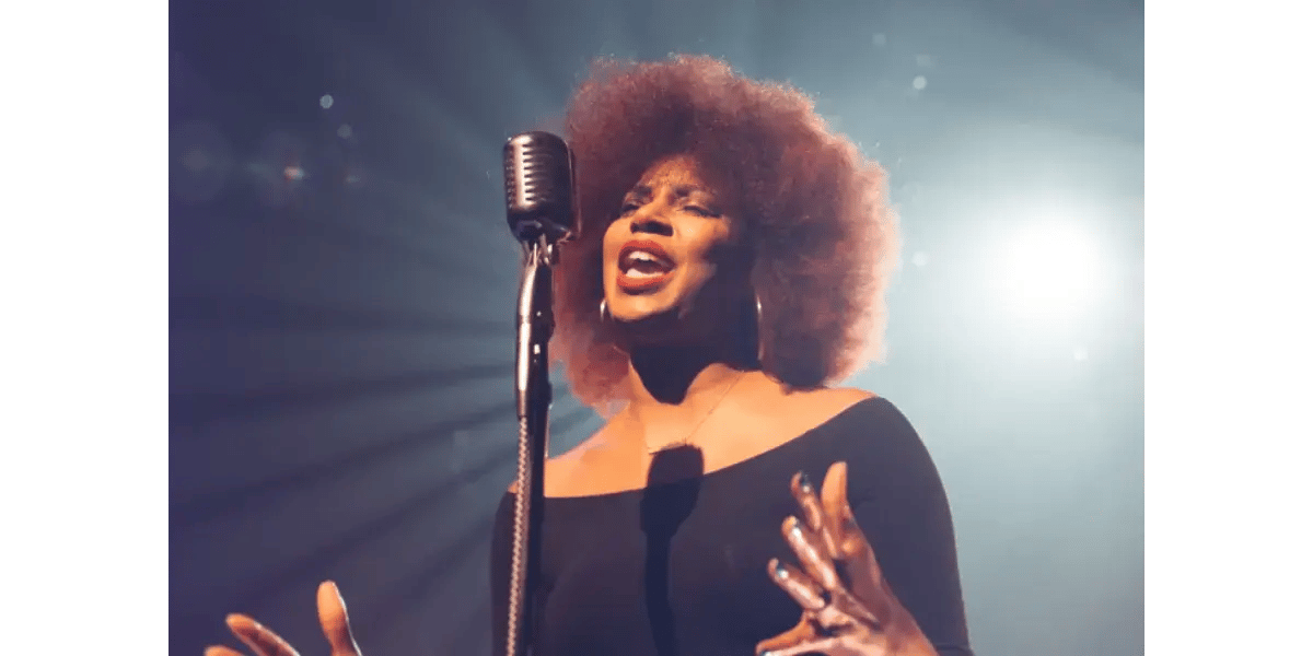 A woman with an afro hairstyle and off-the-shoulder dress singing with an expressive smile into a classic silver microphone on a dark stage with warm bokeh lights.