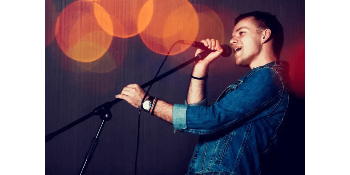 Male singer in a blue shirt performing live on stage with a microphone, representing vocal stretch and muscle balance.