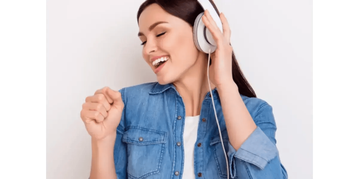 A smiling young woman with light brown hair wearing over-ear white headphones, eyes closed and happily singing.