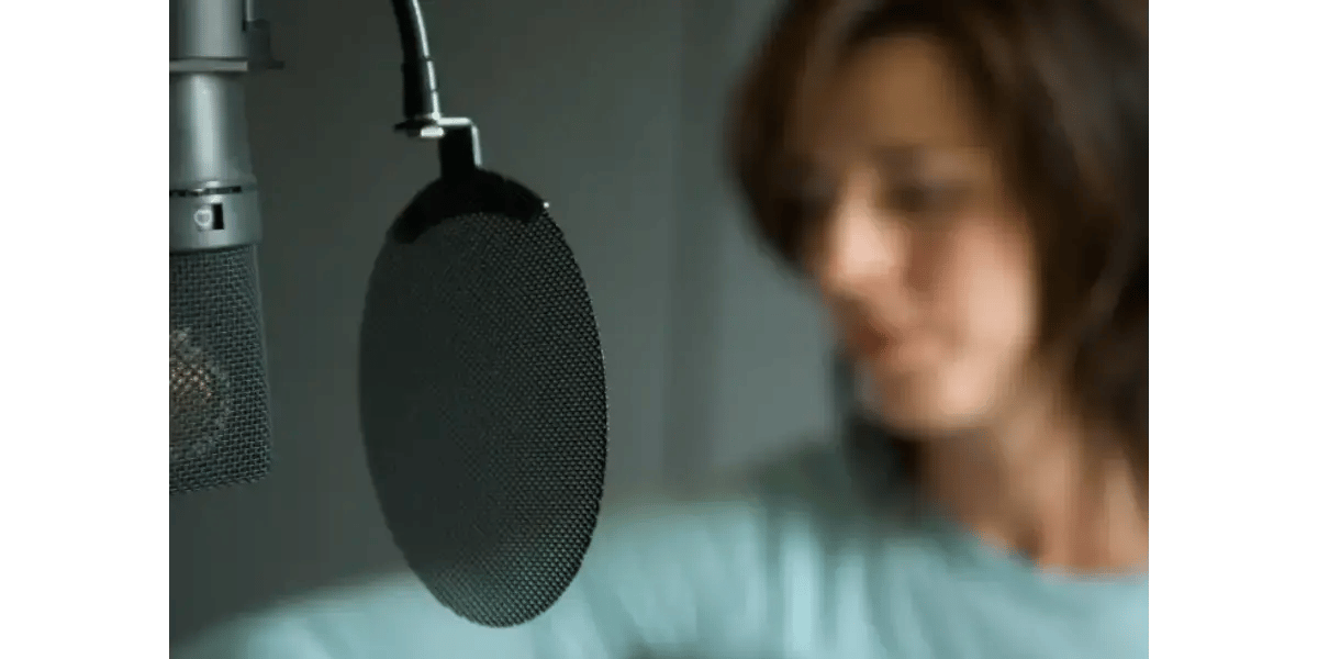 A close-up of a studio microphone with a round pop filter, with a singer slightly out of focus in the background.