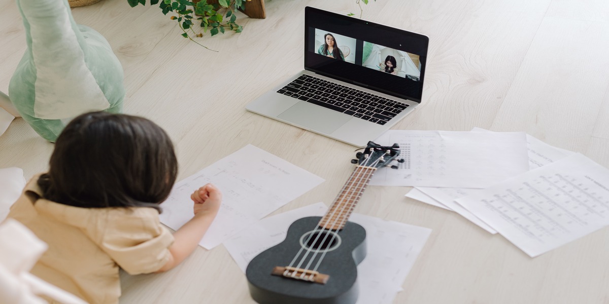 Child participating in an online voice lesson on a laptop next to a ukulele and sheet music.