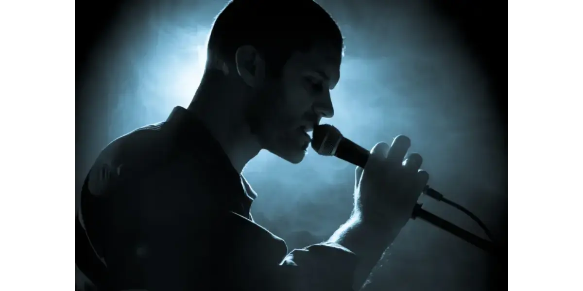 A male vocalist in profile, singing into a microphone in a dark, blue-lit studio setting