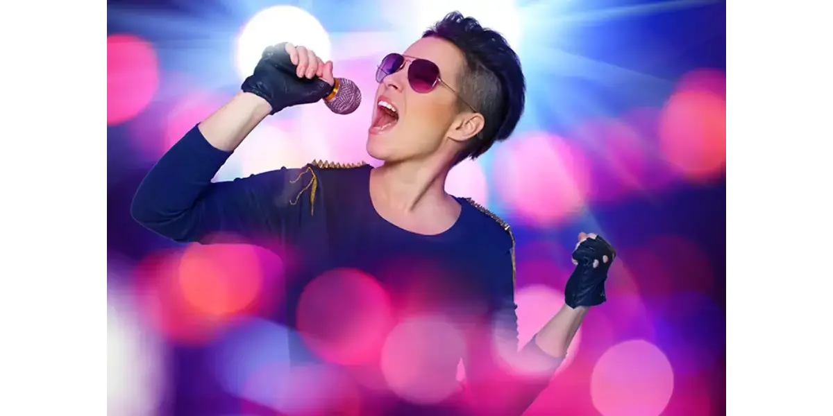 A stylish female vocalist wearing sunglasses and fingerless gloves holding a microphone close to her mouth while singing on stage, illuminated by vibrant pink, blue, and purple bokeh background lights.