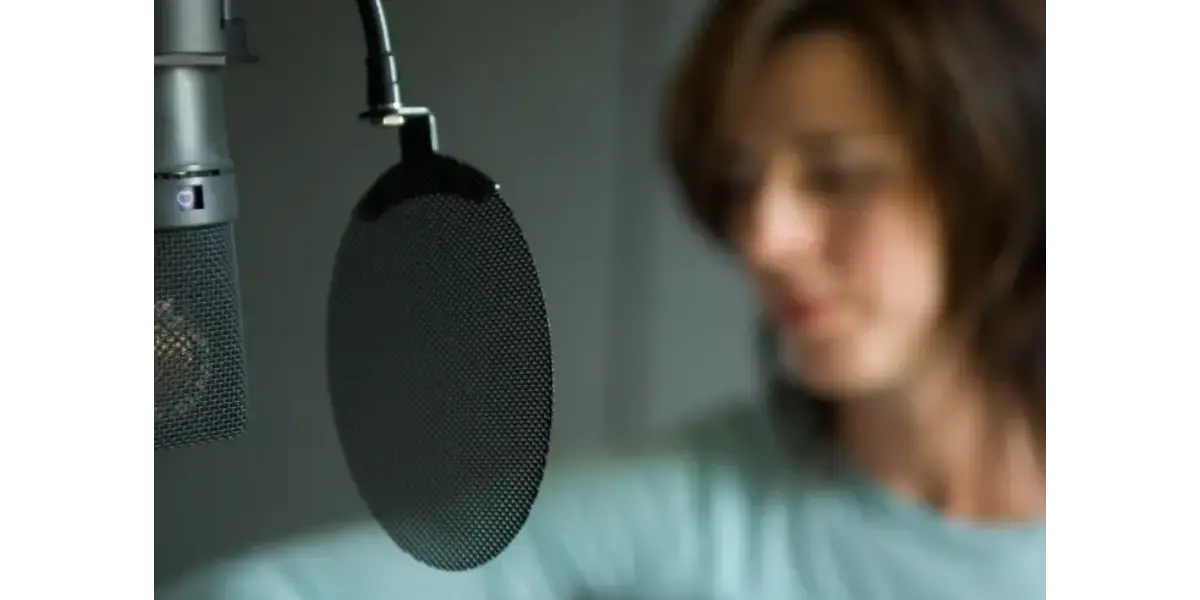 A close-up of a studio microphone with a round pop filter, with a singer slightly out of focus in the background.